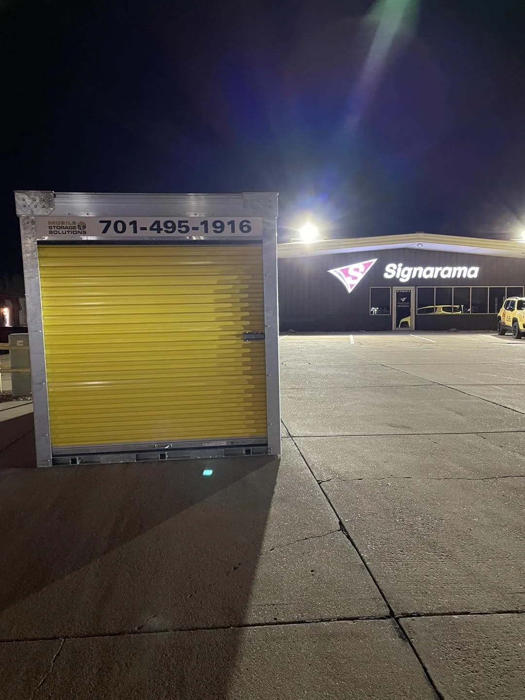 A yellow storage unit with a roller door is placed in front of a parking lot at night. In the background, there is a sign for Signarama, a printing service business, on a dark-colored building, with a couple of parked vehicles nearby.