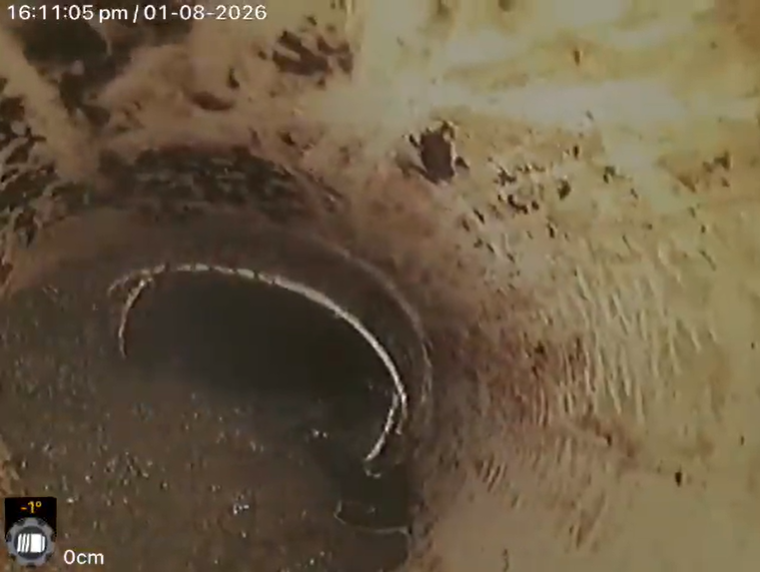 Close-up view of a pipe in a dirt or concrete excavation site, with a timestamp and temperature display on the top left corner and a depth indicator at the bottom left corner.