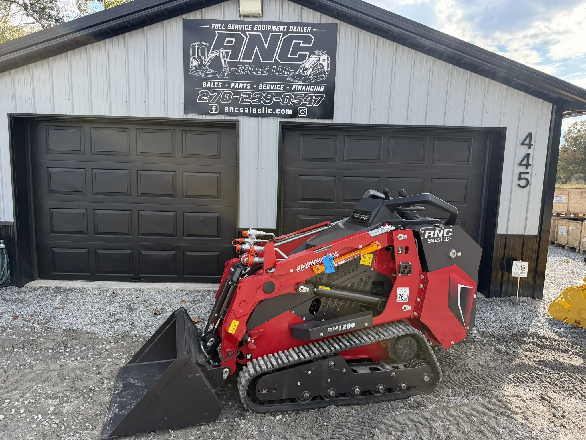 A small red and black mini excavator with tracks, resting in front of a building with a sign that reads "ANC Sales LLC" offering full service equipment sales, parts, service, and financing.  Rhinoceros RH1200 Mini Skid Steer