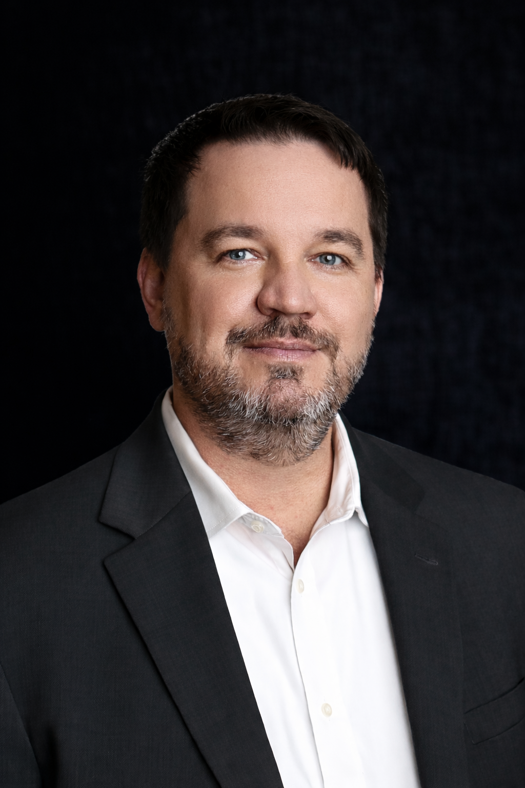 A man with dark hair and a beard wearing a black blazer and white shirt, smiling against a dark background.