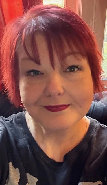 A woman with short red hair and dark lipstick smiling for a selfie indoors.