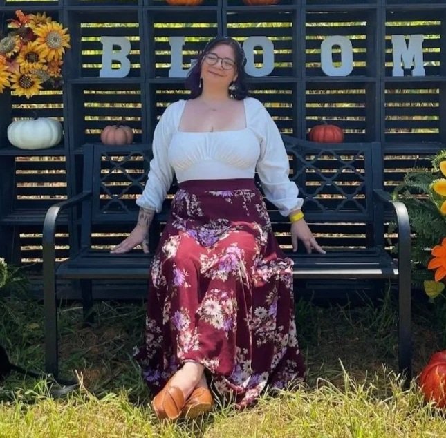 A woman sitting on a black park bench with pumpkins and sunflowers around her. Behind her, a black slatted backdrop with greenery. She is smiling, wearing a white blouse and a long, floral maroon skirt, with brown shoes and a yellow bracelet.