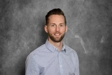 A man with short brown hair and a beard, wearing a blue and white striped dress shirt, smiling at the camera against a gray textured background.