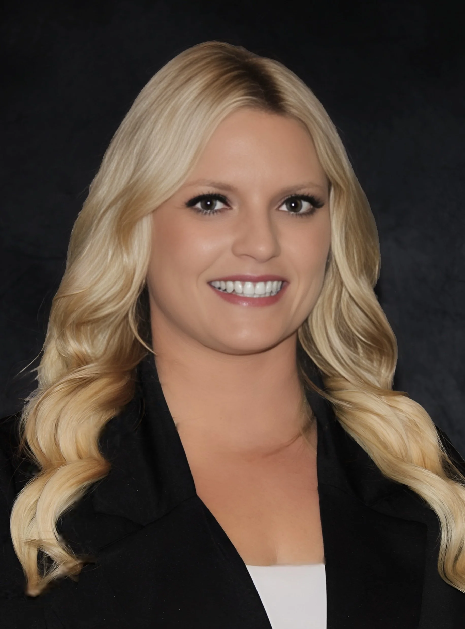 A professional woman with long blonde hair styled in loose waves, wearing a black blazer and smiling against a dark background.