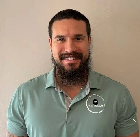 A man with short dark hair, a beard, and a mustache smiling and wearing a light green polo shirt with a logo that says 'HEALING COUNSELING' on the chest, standing against a plain beige wall.