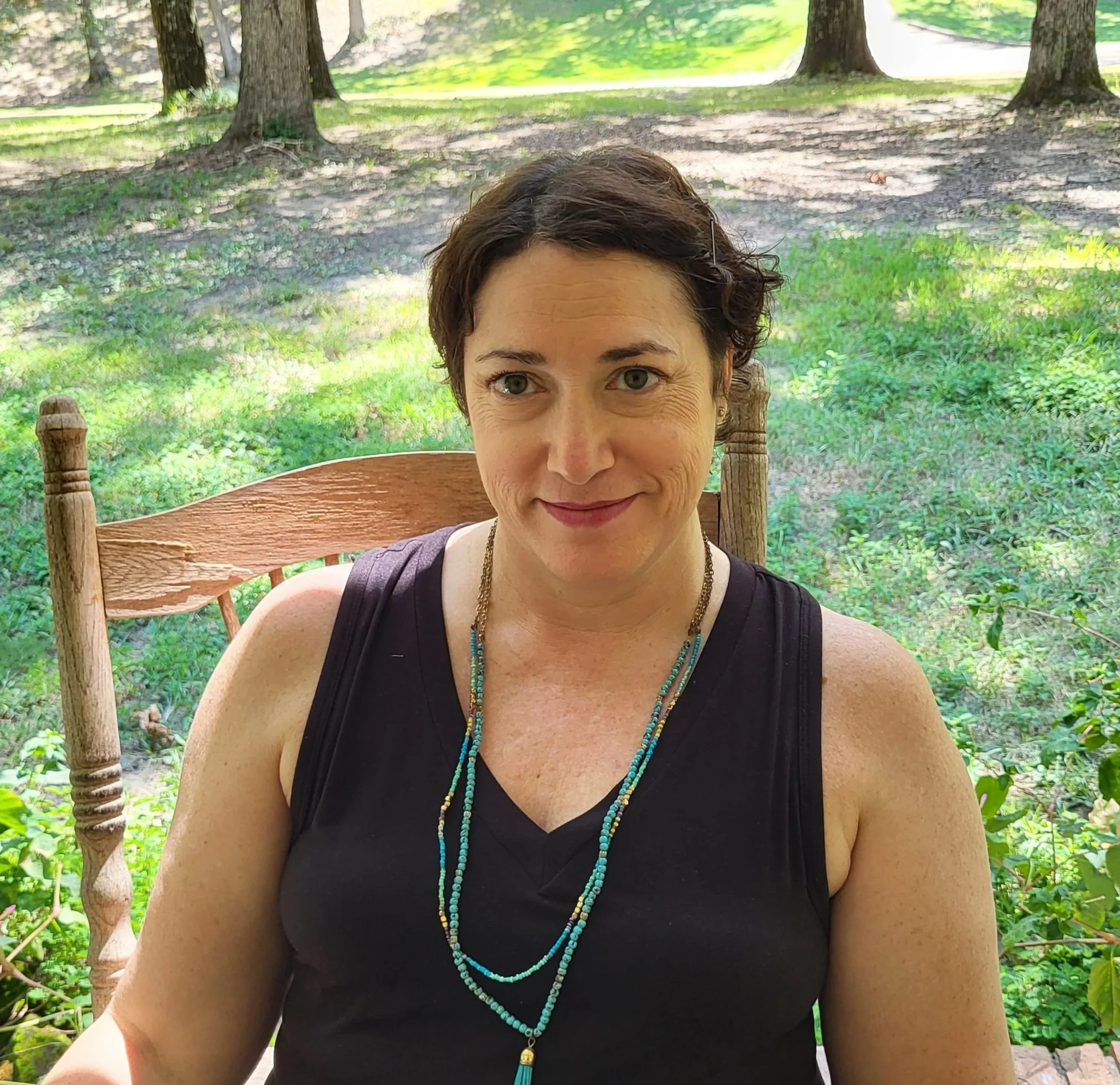 A woman with short dark hair sits on a wooden chair outdoors in a park, smiling gently, wearing a black sleeveless top and turquoise beaded necklaces, with trees and green grass in the background.
