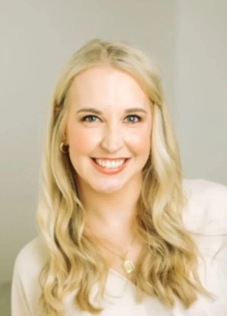 A smiling woman with long, wavy blonde hair, wearing a white top and a necklace, standing against a neutral background.