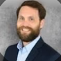 A headshot of a man with a beard, wearing a dark blazer and light blue shirt, smiling against a neutral background.
