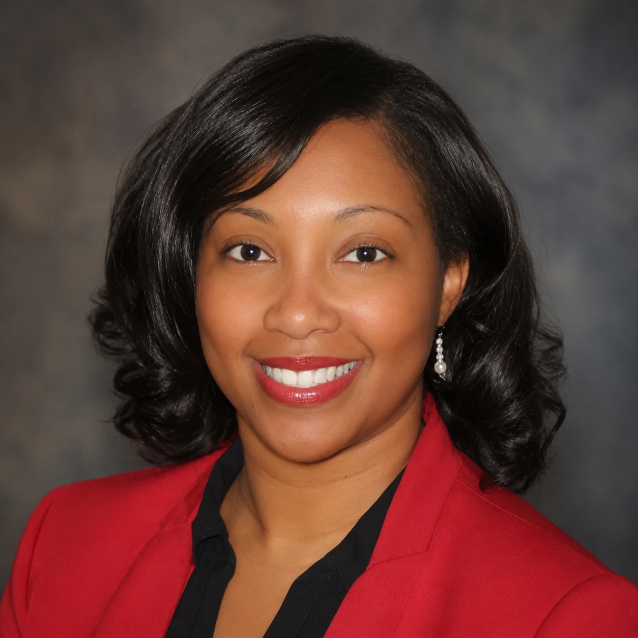 Image of a smiling woman with dark hair, wearing a red blazer and black shirt, with earrings, against a dark background.