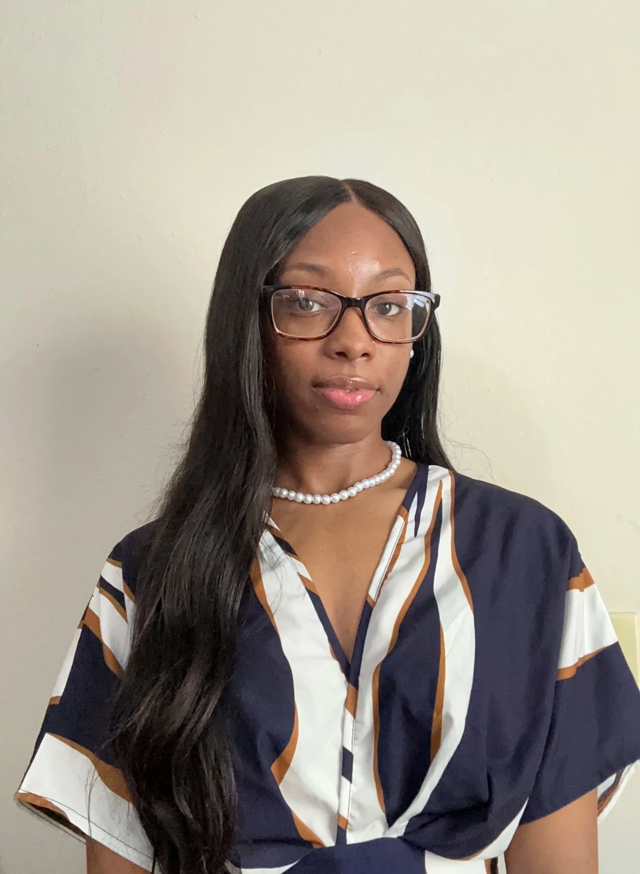 A woman with long black hair, wearing glasses, a pearl necklace, and a patterned blouse, standing against a plain cream-colored wall.