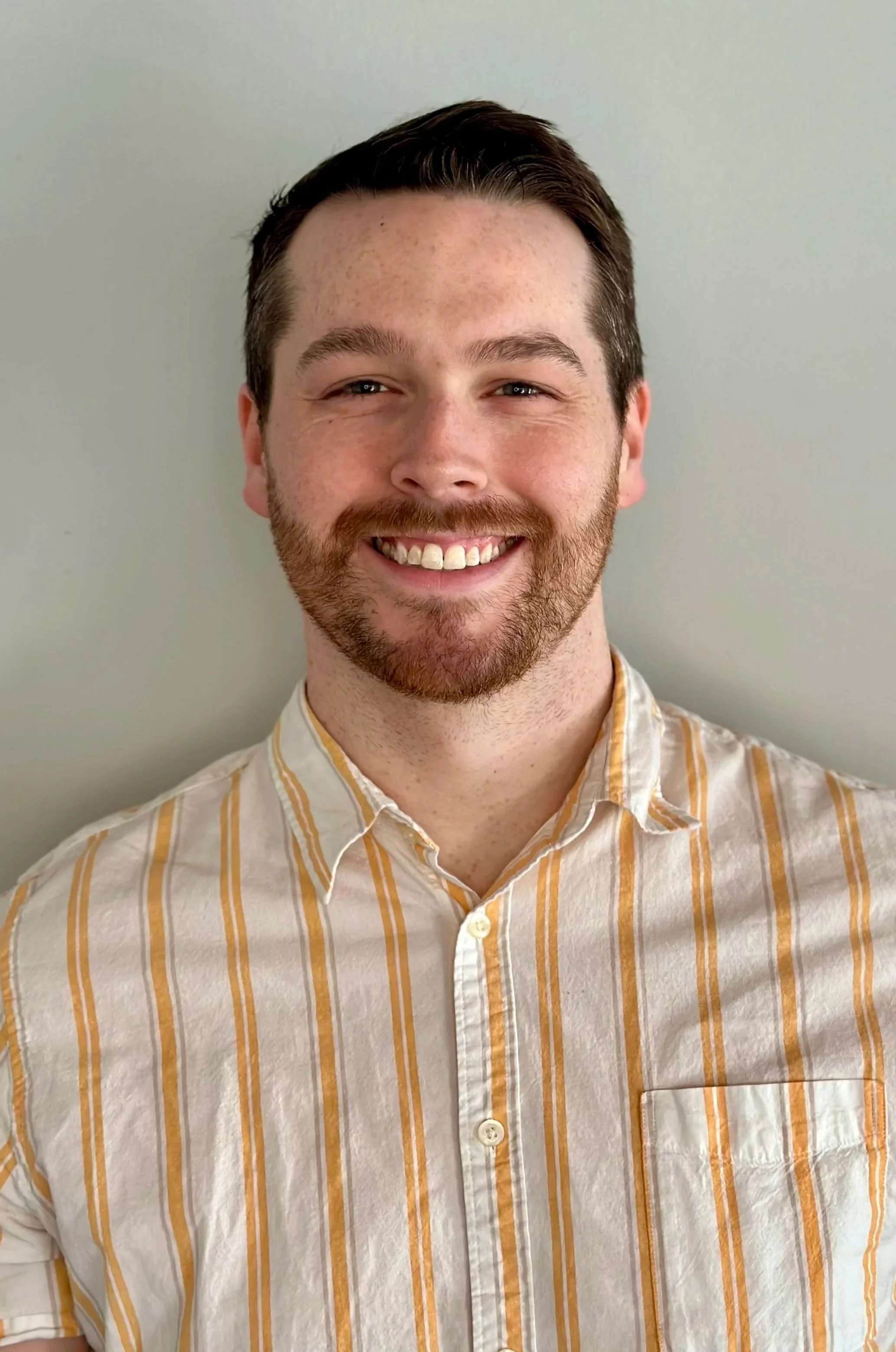 A young man with brown hair, a beard, and a mustache smiling, wearing a yellow and white striped button-up shirt, standing against a plain light-colored wall.