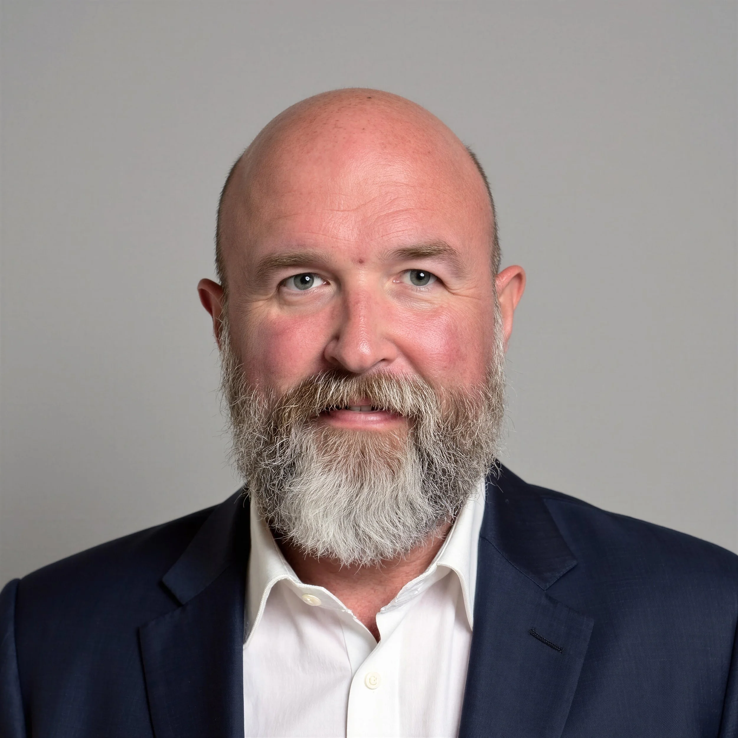 A headshot of a smiling man, bald with a distinguished gray beard, wearing a dark blazer and white collared shirt, positioned against a plain gray background.