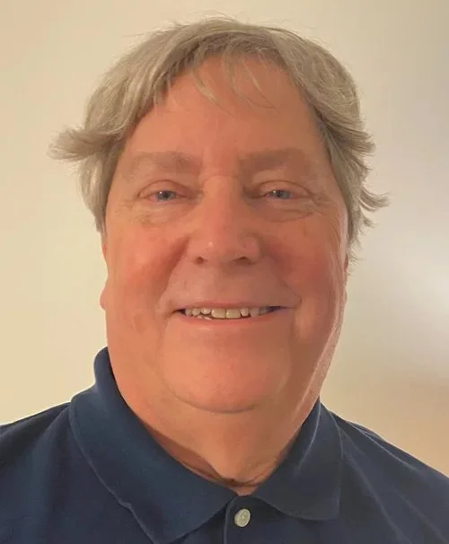 Image of a smiling middle-aged man with gray hair wearing a dark blue collared shirt, standing against a plain light-colored background.