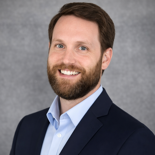 A headshot of a man with a beard, wearing a dark blazer and light blue shirt, smiling against a neutral background.