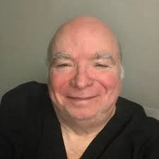 Close-up of a smiling bald man with a mustache, wearing a black shirt, against a light green background.