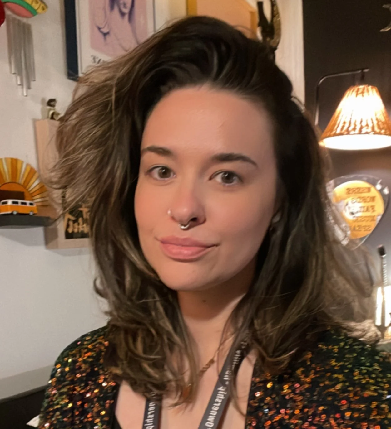 A headshot of a woman with brown wavy hair and a septum piercing, wearing a black top.
