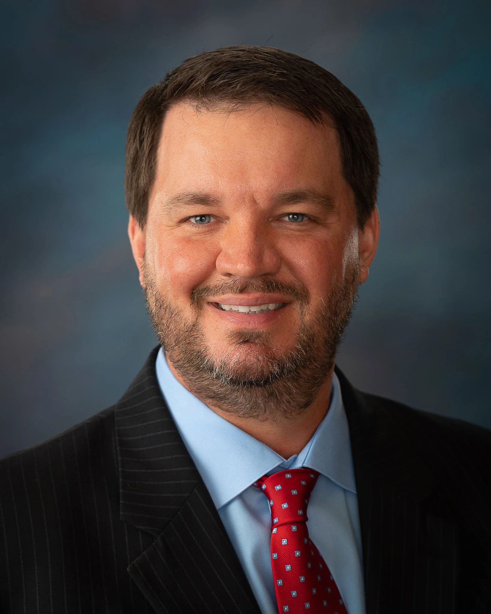 A man with dark hair and a beard wearing a black blazer and white shirt, smiling against a dark background.