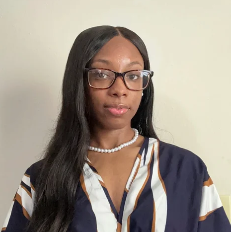 Image of a woman with long black hair, glasses, and a pearl necklace, wearing a patterned blouse, standing against a plain light-colored wall.