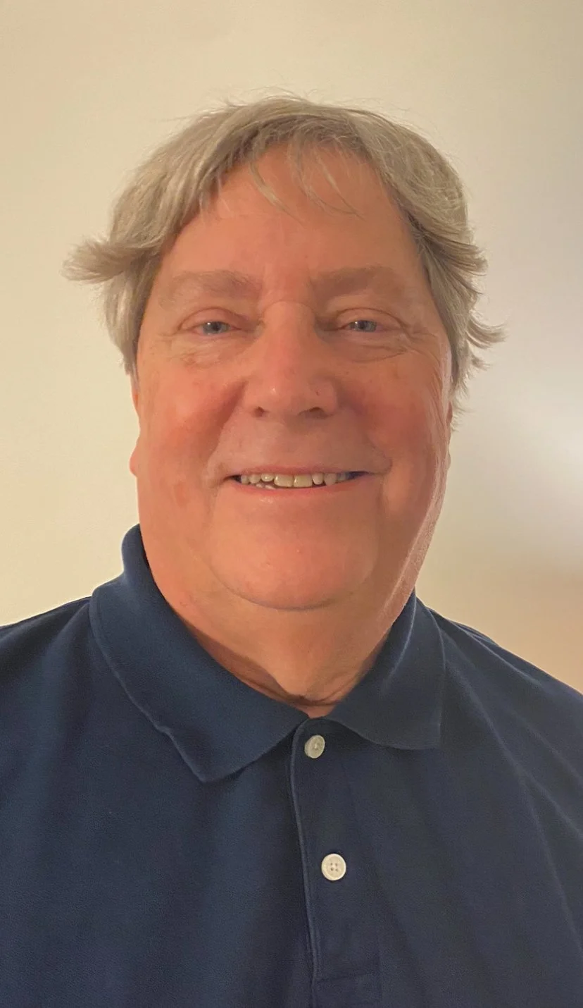 A smiling older man with gray hair wearing a dark blue collared shirt, standing against a plain, light-colored background.