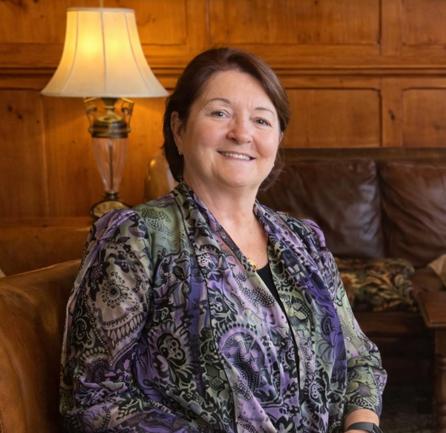 A smiling middle-aged woman with short brown hair, wearing a colorful paisley blouse, sitting on a leather couch in a warmly lit room with wooden paneling and a table lamp in the background.
