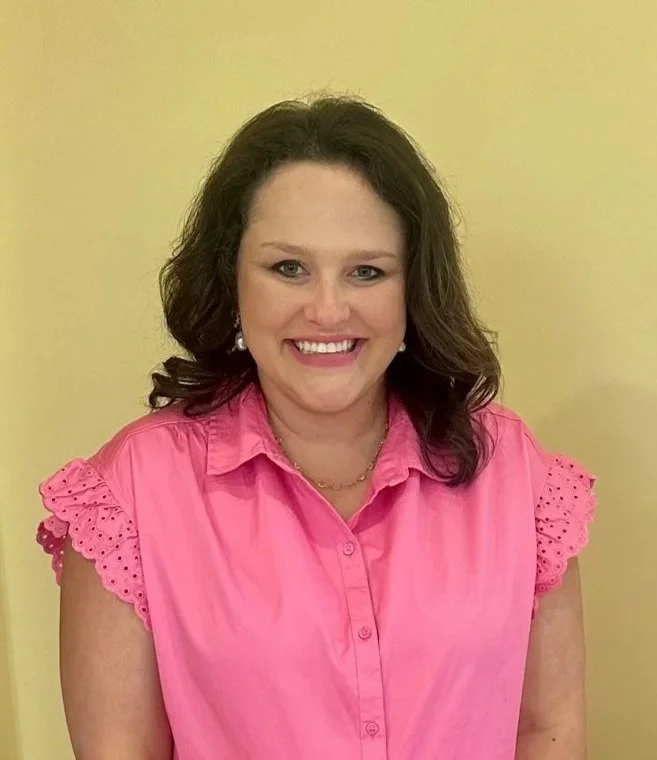 Smiling woman with wavy brown hair wearing a pink sleeveless blouse and pearl earrings against a yellow background.