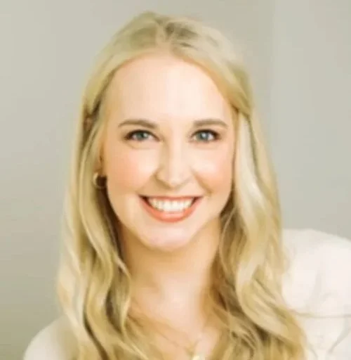 A smiling woman with long, wavy blonde hair, wearing a white top and a necklace, standing against a neutral background.