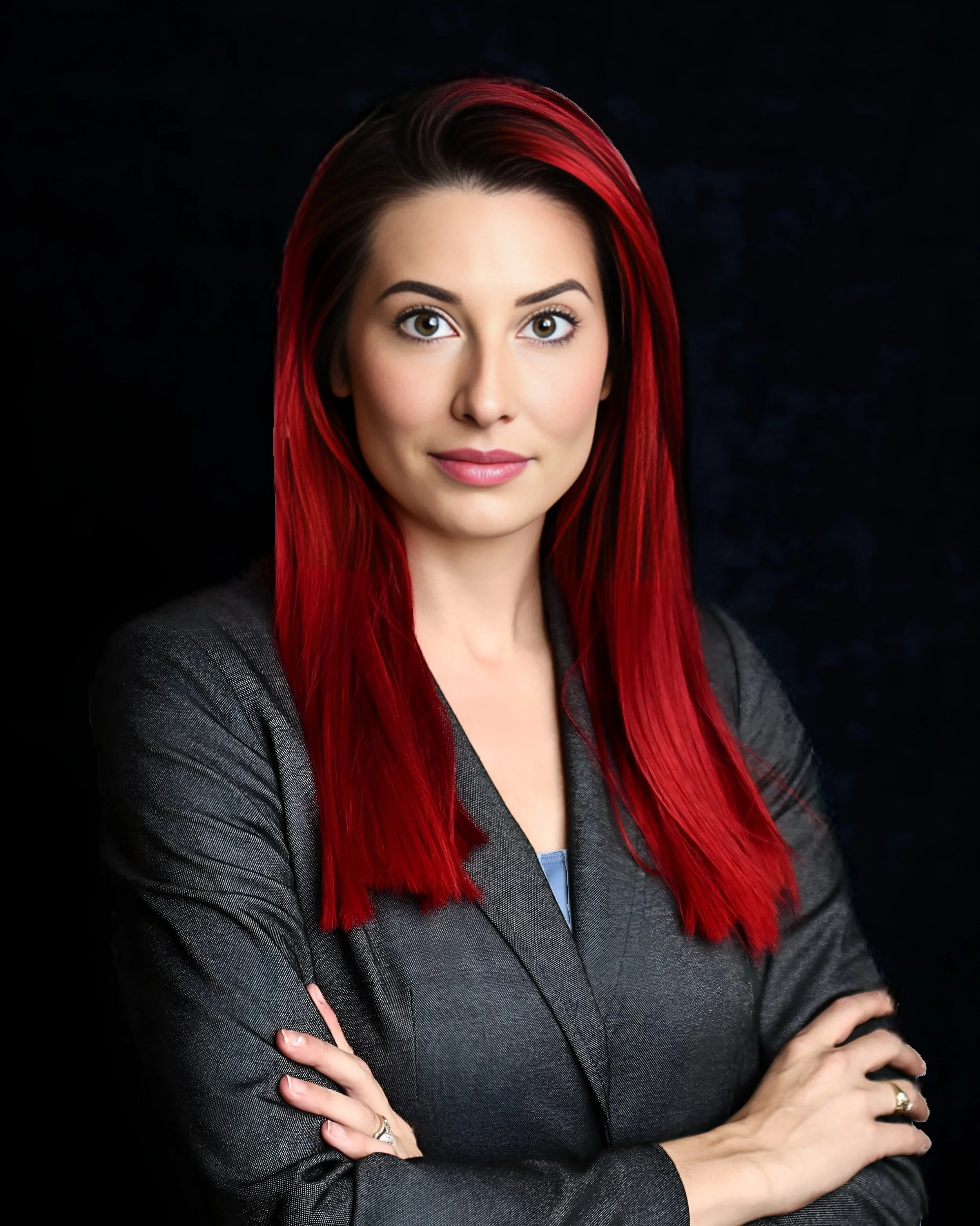 A woman with long red hair, wearing a dark blazer, standing with arms crossed against a dark background.