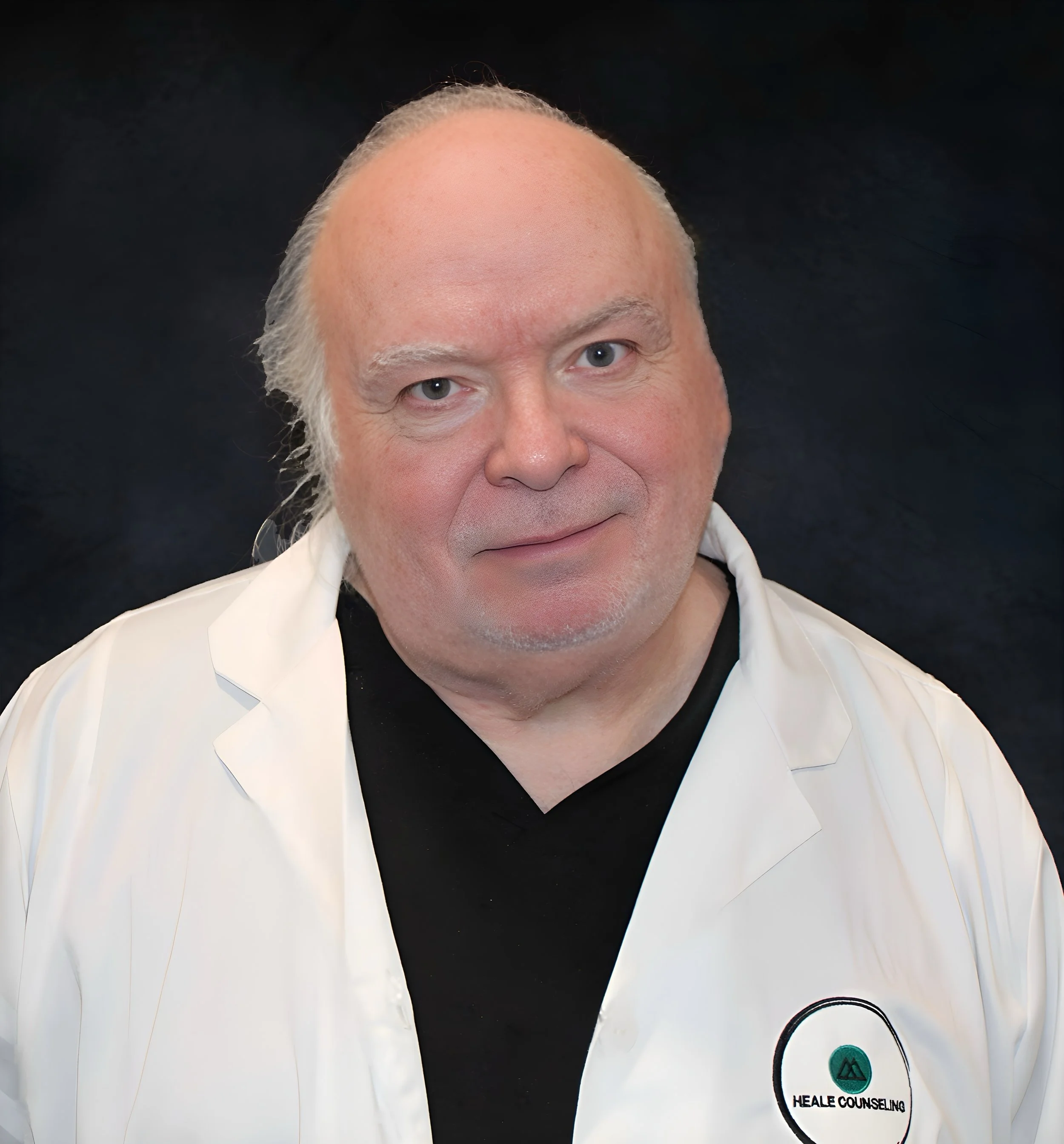 A middle-aged man with light skin, white hair, and a friendly expression, wearing a white jacket with 'Heale Counseling' logo over a black shirt, against a dark background.