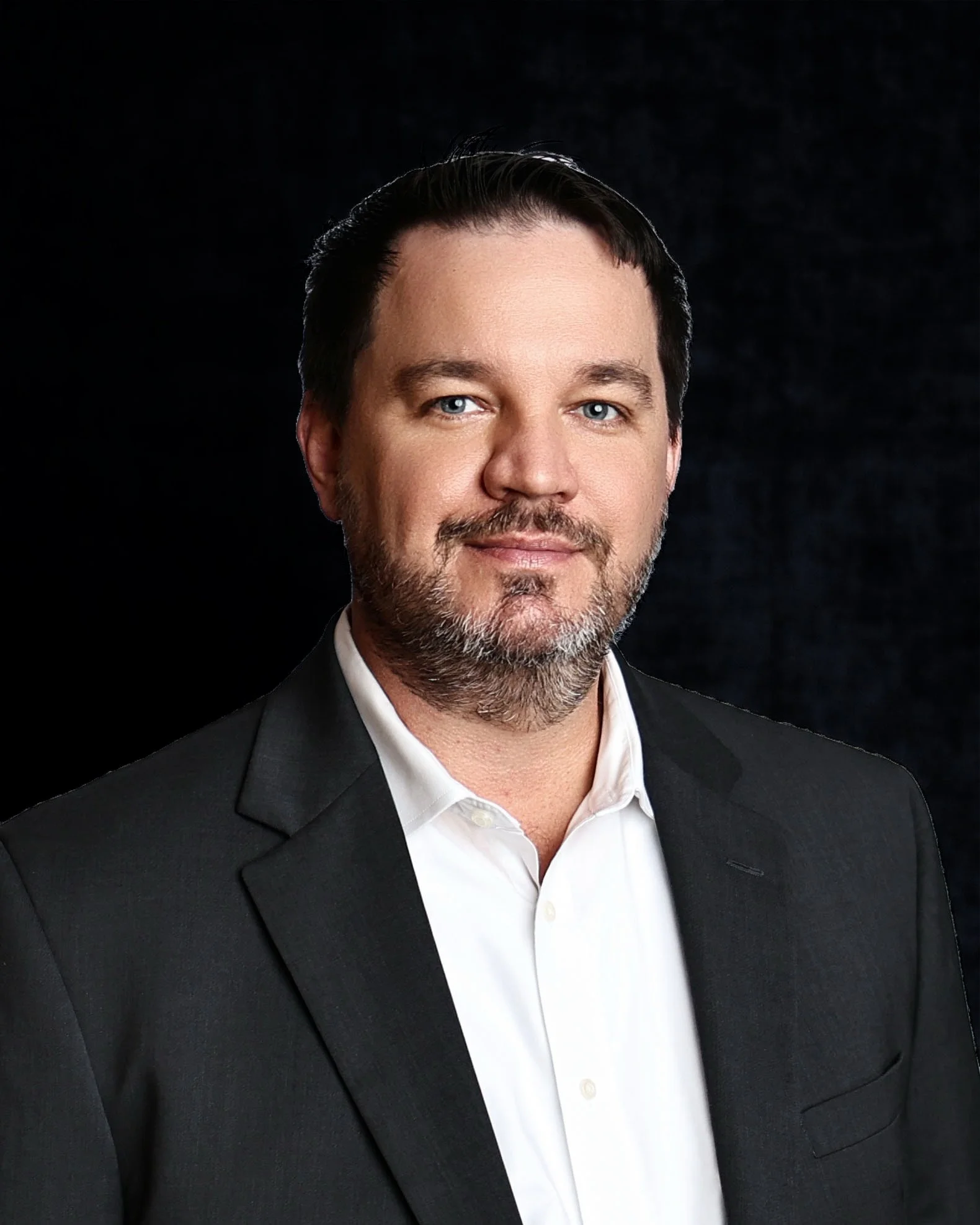 A man with dark hair and a beard wearing a black blazer and white shirt, smiling against a dark background.