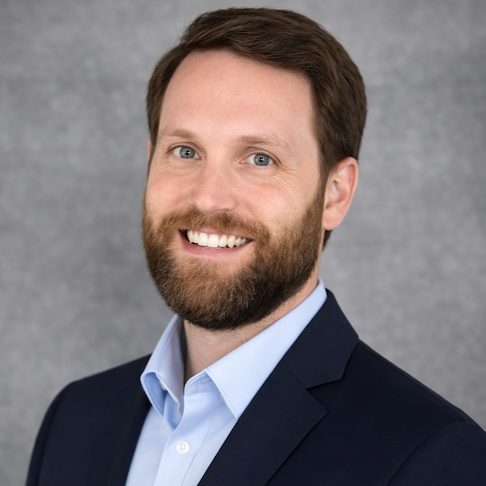 A headshot of a man with a beard, wearing a dark blazer and light blue shirt, smiling against a neutral background.