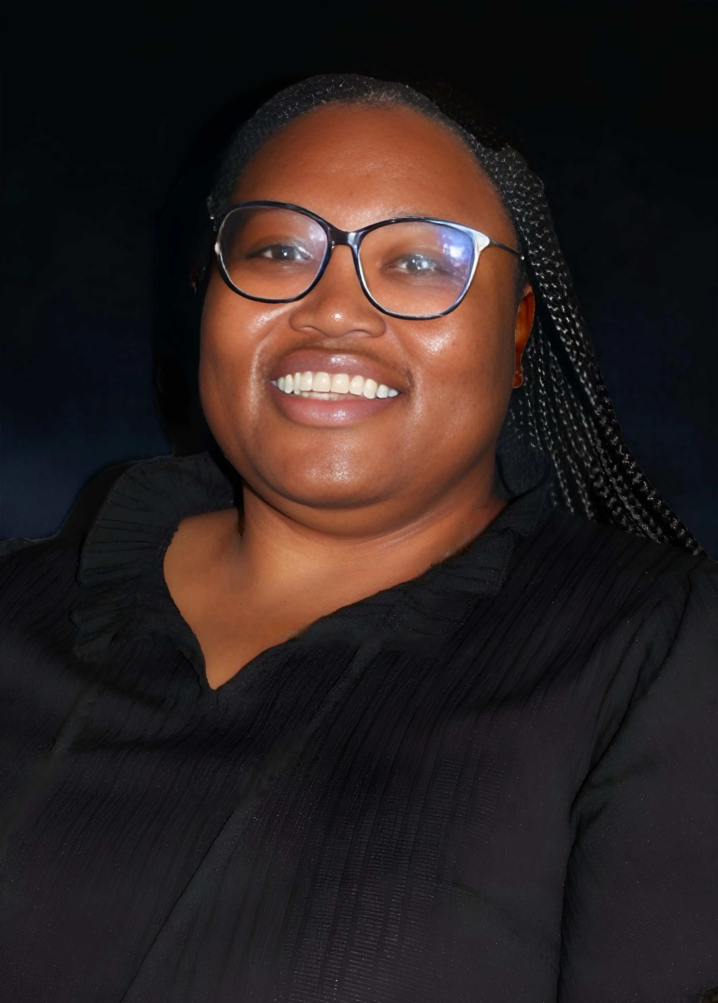 A woman smiling, wearing glasses, with black hair styled in braids, dressed in a black top, against a dark background.