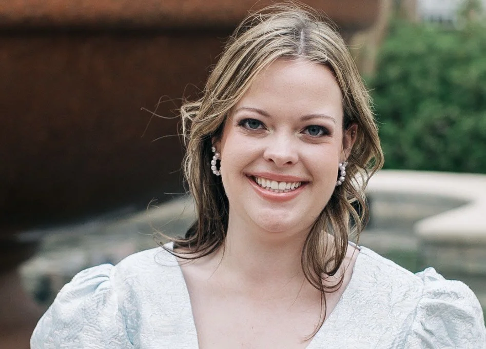 A woman with light brown, wavy hair smiling outdoors, wearing pearl earrings and a white top.