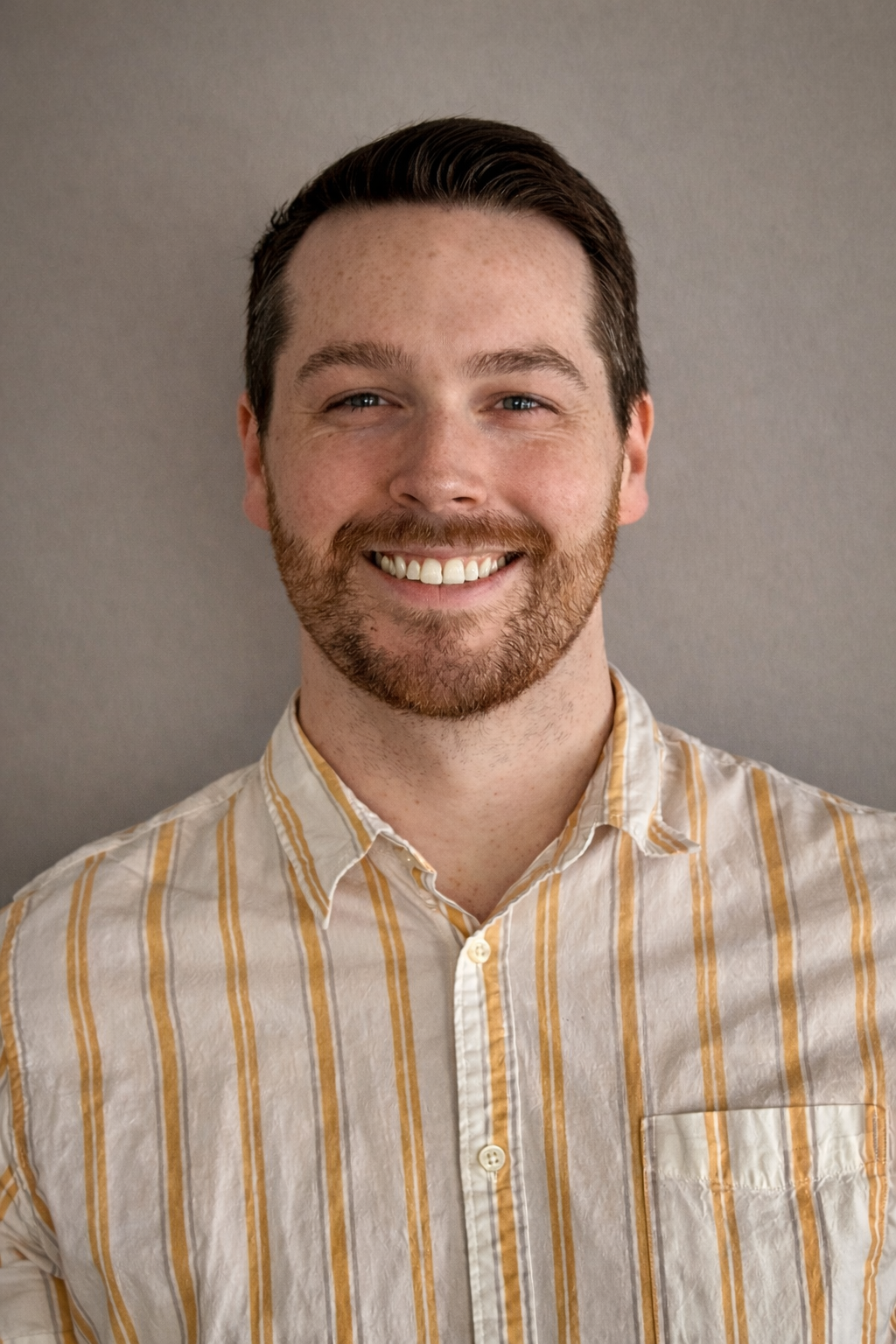 A young man with brown hair, a beard, and a mustache smiling, wearing a yellow and white striped button-up shirt, standing against a plain light-colored wall.