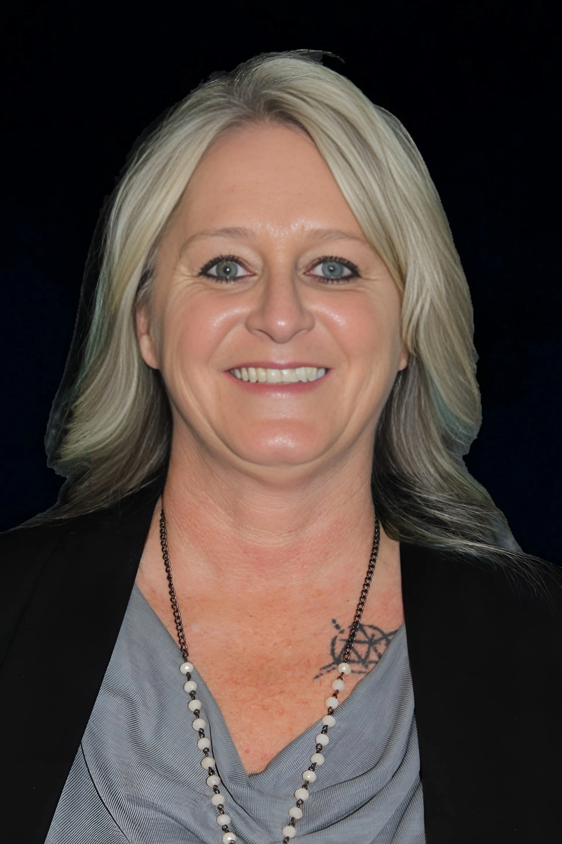 A woman with blonde hair smiling, wearing a black blazer, a gray top, and a pearl necklace, with a tattoo visible on her chest, against a dark background.