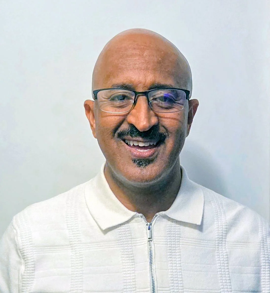 Smiling man with glasses, bald head, mustache, and goatee, wearing a white zip-up jacket, standing against a plain white background.