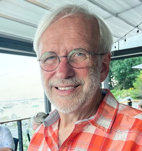 Smiling elderly man with white hair, glasses, and a beard wearing an orange plaid shirt, indoors with windows showing an outdoor view.