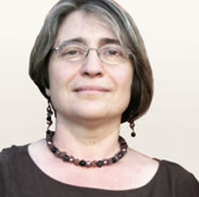 A woman with short brown hair, glasses, and earrings, smiling slightly, wearing a black top and a beaded necklace and earrings.