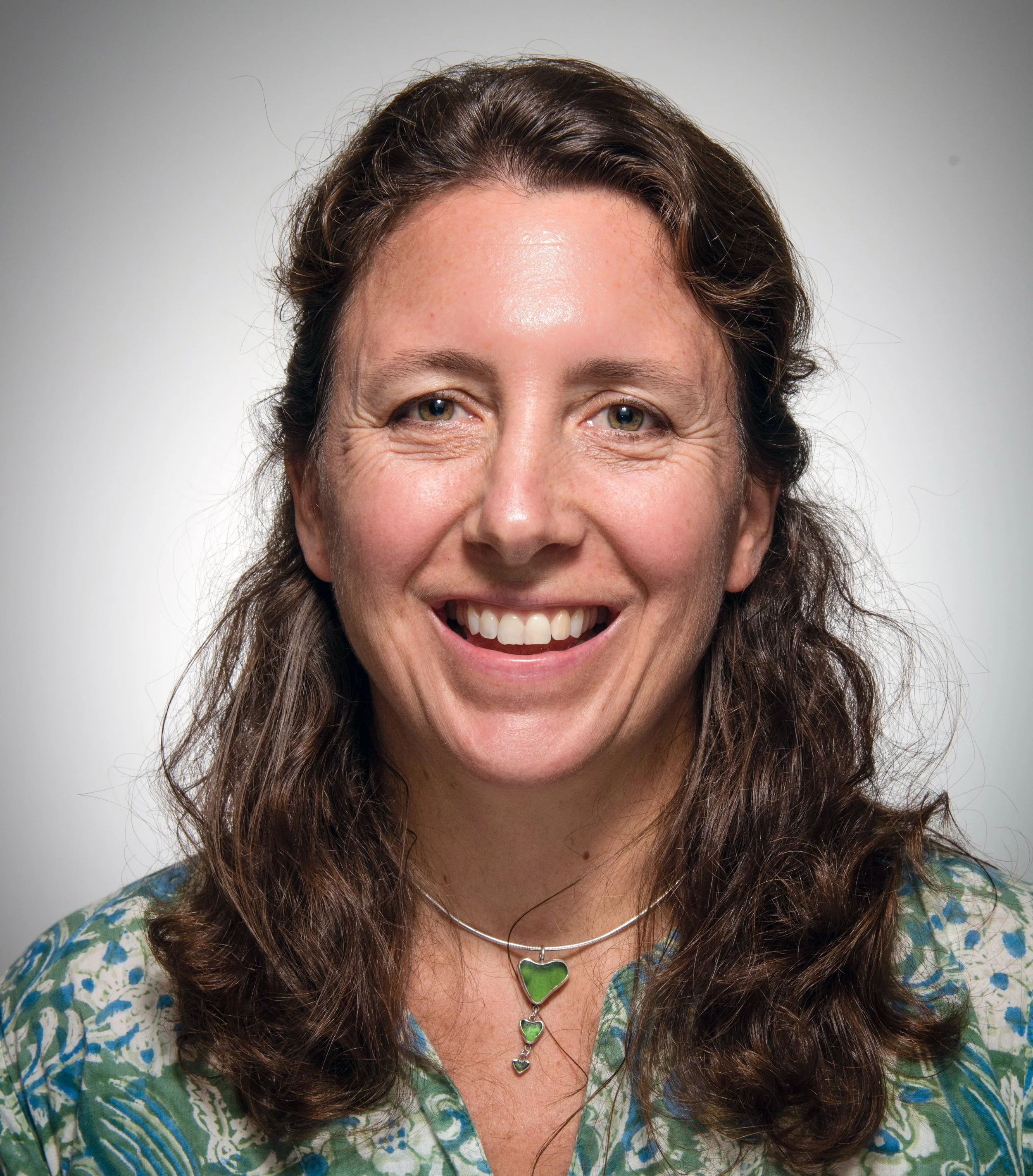 Close-up of a smiling woman with curly brown hair, wearing a green and blue patterned shirt and a silver necklace with a large green heart pendant, standing against a plain, light gray background.