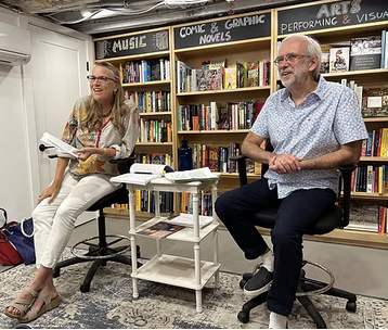 Two people sitting on stools in a bookstore or library, with a bookshelf behind them filled with books. They appear to be having a discussion or presentation.