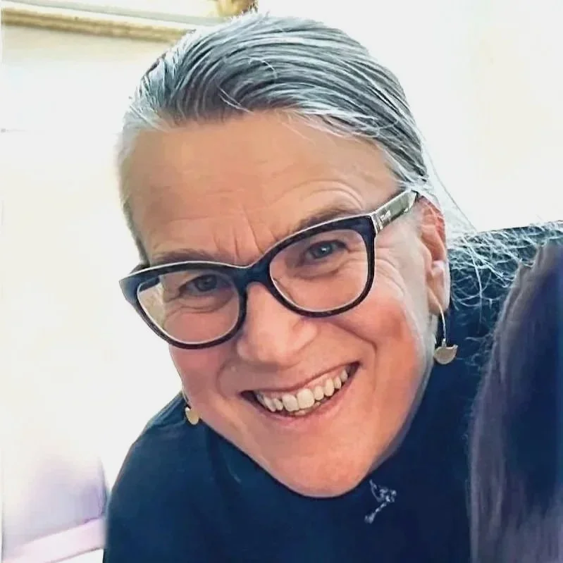 Close-up of a smiling woman with gray hair, wearing glasses and earrings.