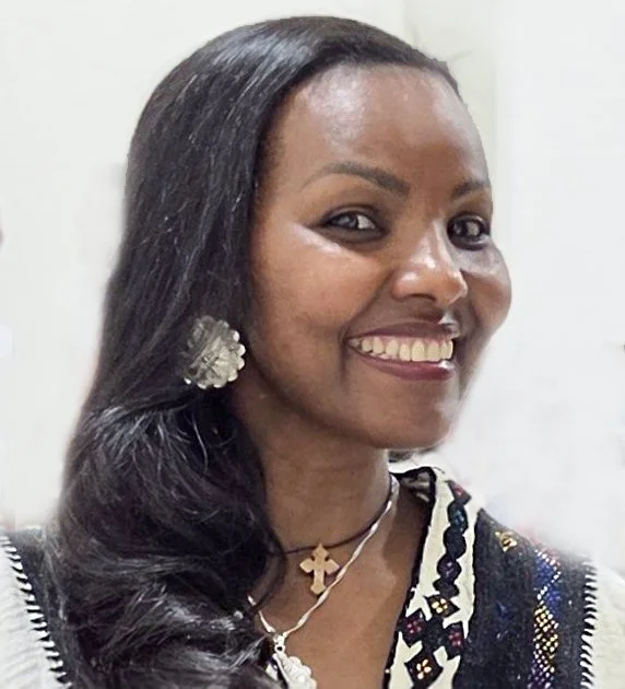 A smiling woman with long black hair wearing large floral earrings, layered necklaces including a cross pendant, and a black and white patterned top.