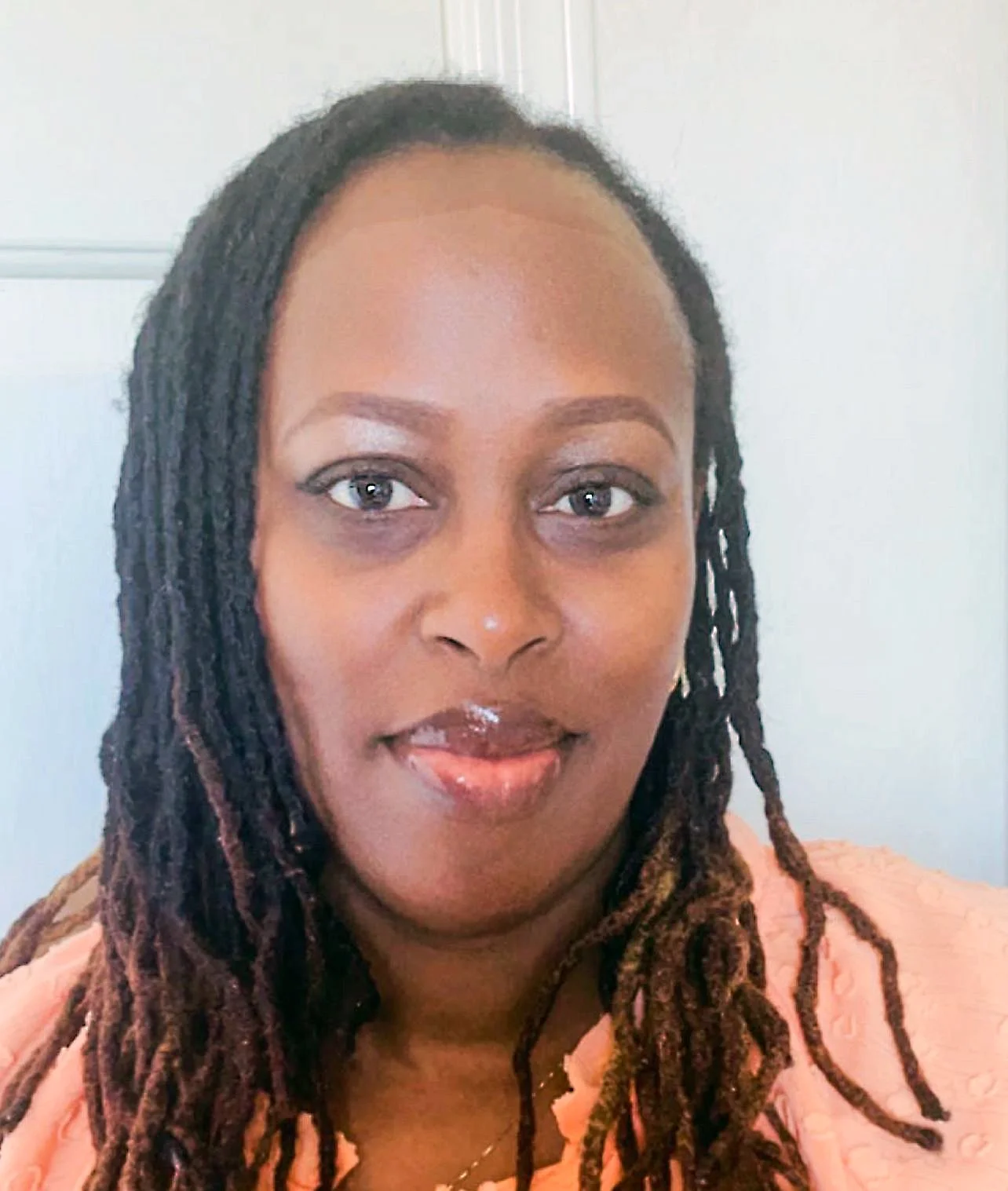 A woman with dark brown skin and long dreadlocks wearing a peach-colored top, looking at the camera with a slight smile.