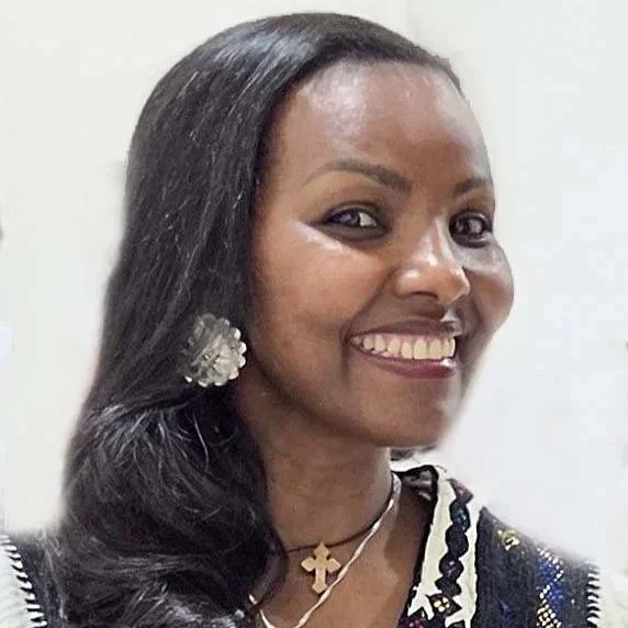 A woman with long, dark hair smiling, wearing large earrings, a necklace with a cross, and a patterned top.