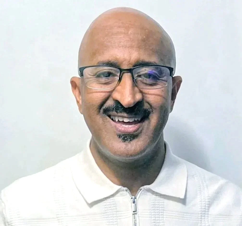 A smiling bald man with glasses, a goatee, and a white zip-up jacket standing against a plain white background.