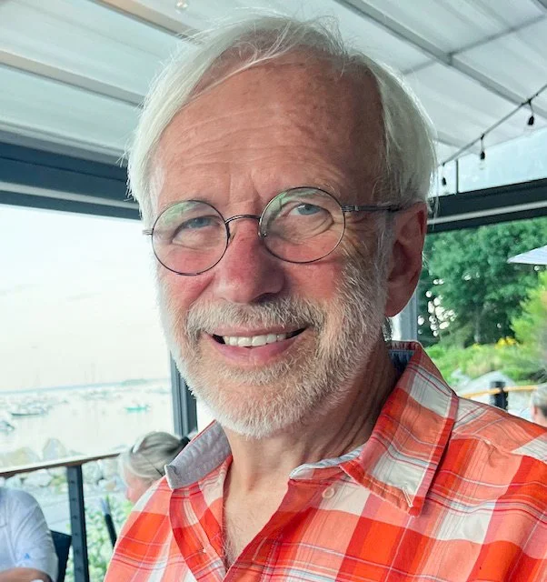 An academic looking man with white hair and glasses, smiling, wearing a red plaid shirt, sitting indoors near a window with a view of boats and water in the background.