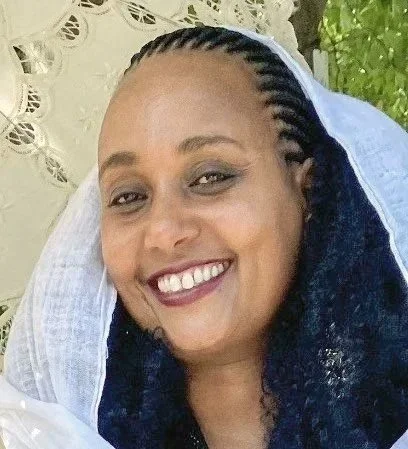 Smiling woman with braids, wearing a headscarf, outdoors with greenery in the background.