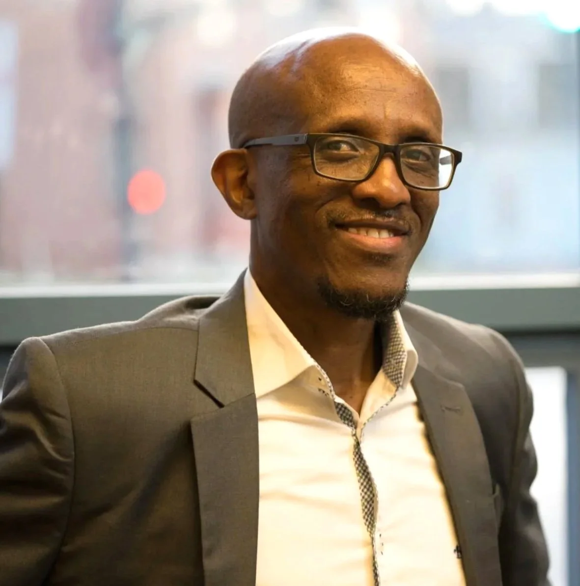 A smiling Rwandan man in a gray suit and glasses, sitting indoors near a window.