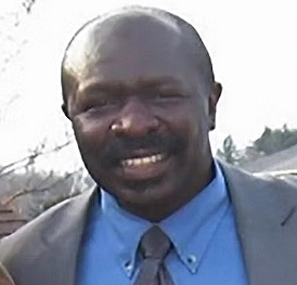 A Rwandan man with a dark complexion, wearing a gray suit, blue shirt, and gray tie, smiling outdoors.