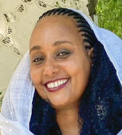 A woman smiling with braided hair outdoors, wearing a white headscarf.