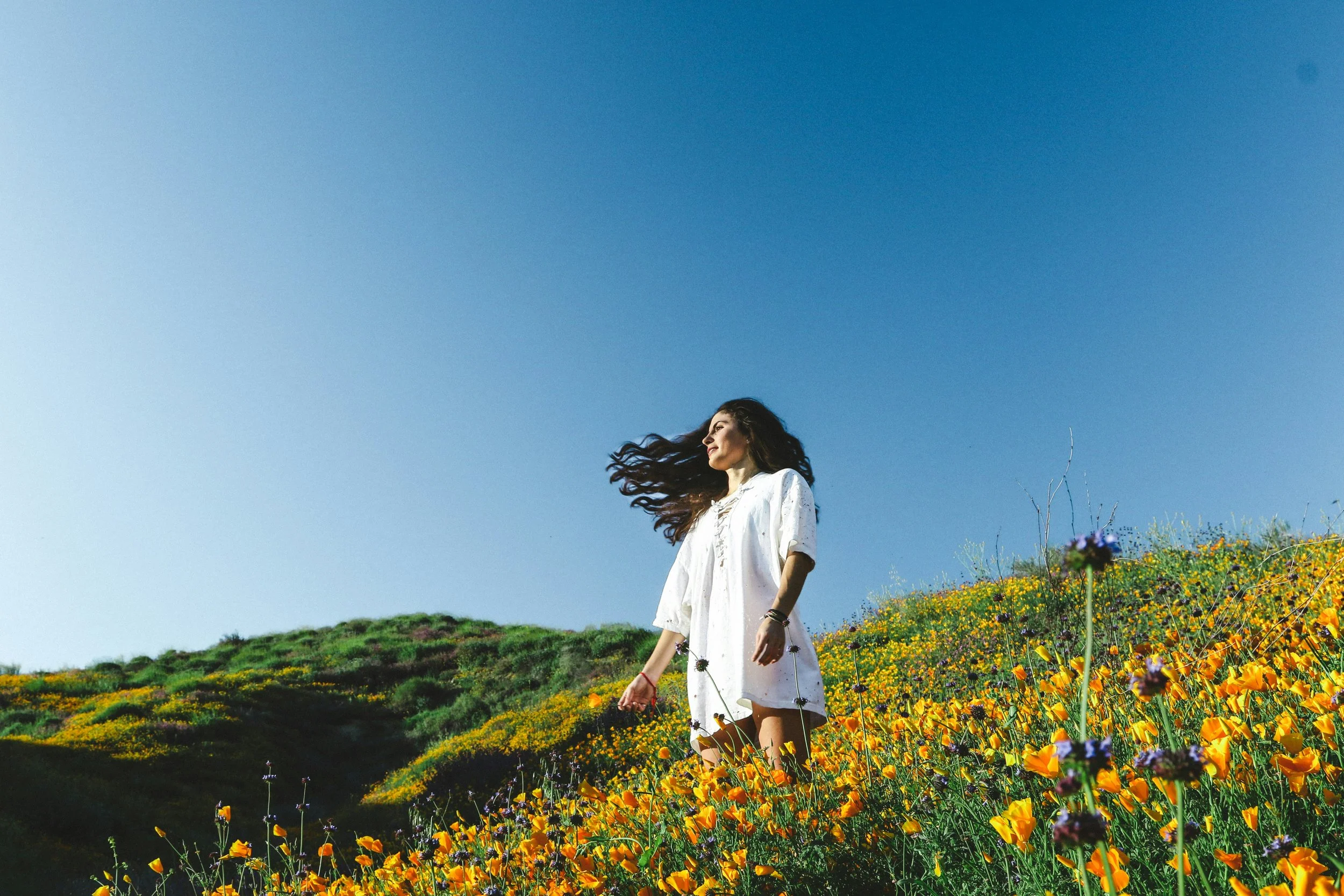 A woman standing in a field of yellow and purple wildflowers on a hillside under a clear blue sky.